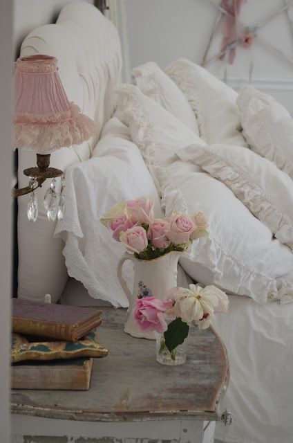 un coin table de nuit en bois ancien portant un vase blanc et des roses à côté d'une tête de lit et des coussins blancs