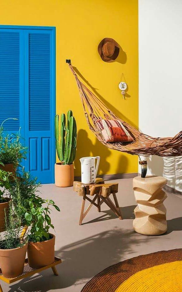 Un espace vibrant avec un mur jaune vif et des volets bleus. Des plantes en pots, un hamac et des éléments en bois