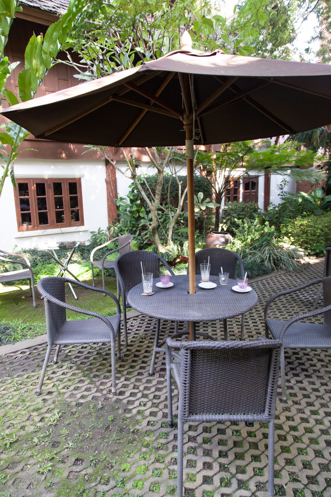table ronde et chaises en osier sous un parasol marron, entourés de verdure et d’une structure blanche en arrière-plan