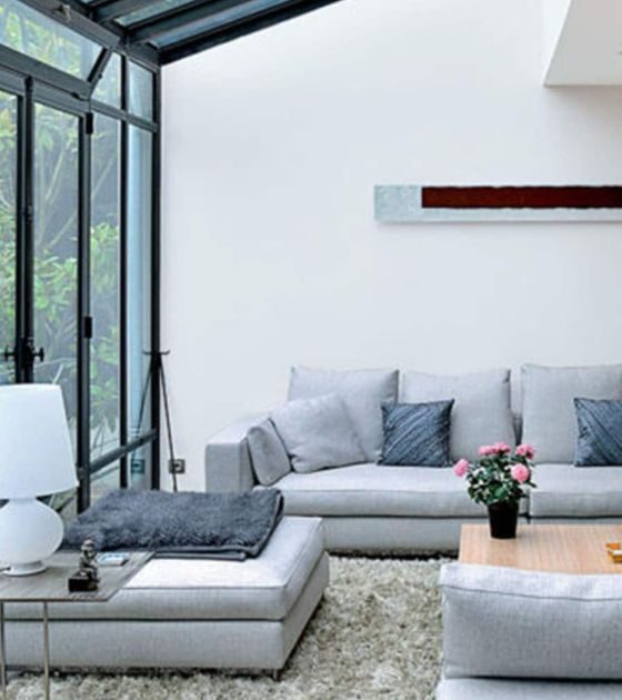 Une véranda moderne avec un canapé en L gris, une table basse en bois, des livres, une décoration florale, et une œuvre d’art murale abstraite.