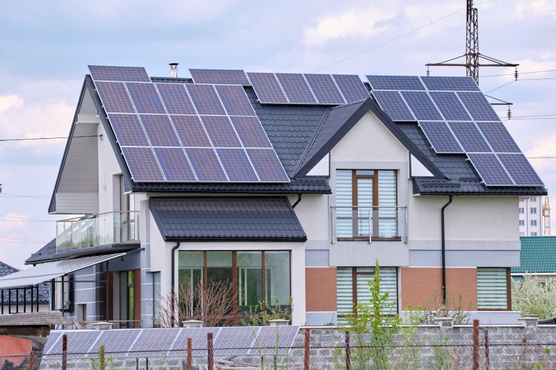 Maison moderne avec toiture en tuiles, équipée de panneaux solaires, jardin verdoyant et balustrade en verre.