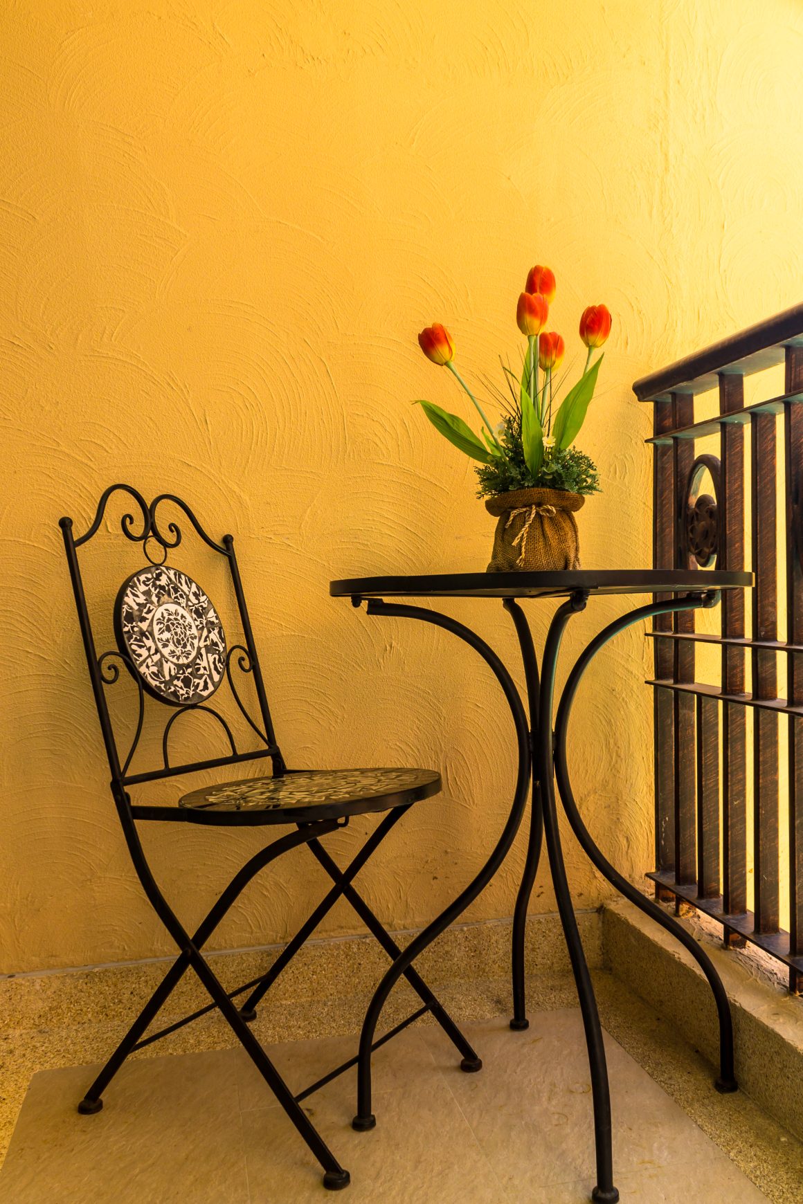 Petit balcon aménagé avec une table ronde en fer, d'une chaise ornée, et d'un pot de tulipes avec un mur en jaune safran.