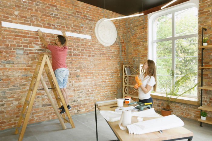 Un homme pose une étagère pendant qu'une femme prépare des outils dans une pièce d'une ancienne maison rénovée.