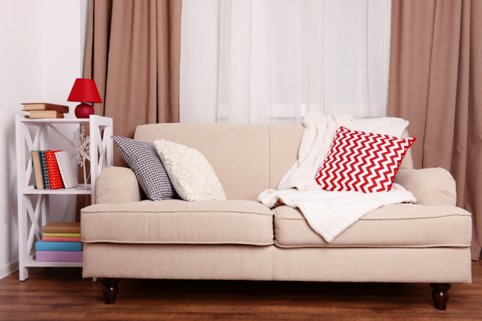 un petit salon avec un canapé 2 places beige, des coussins colorés, une couverture douce, une lampe rouge et une étagère pleine de livres.