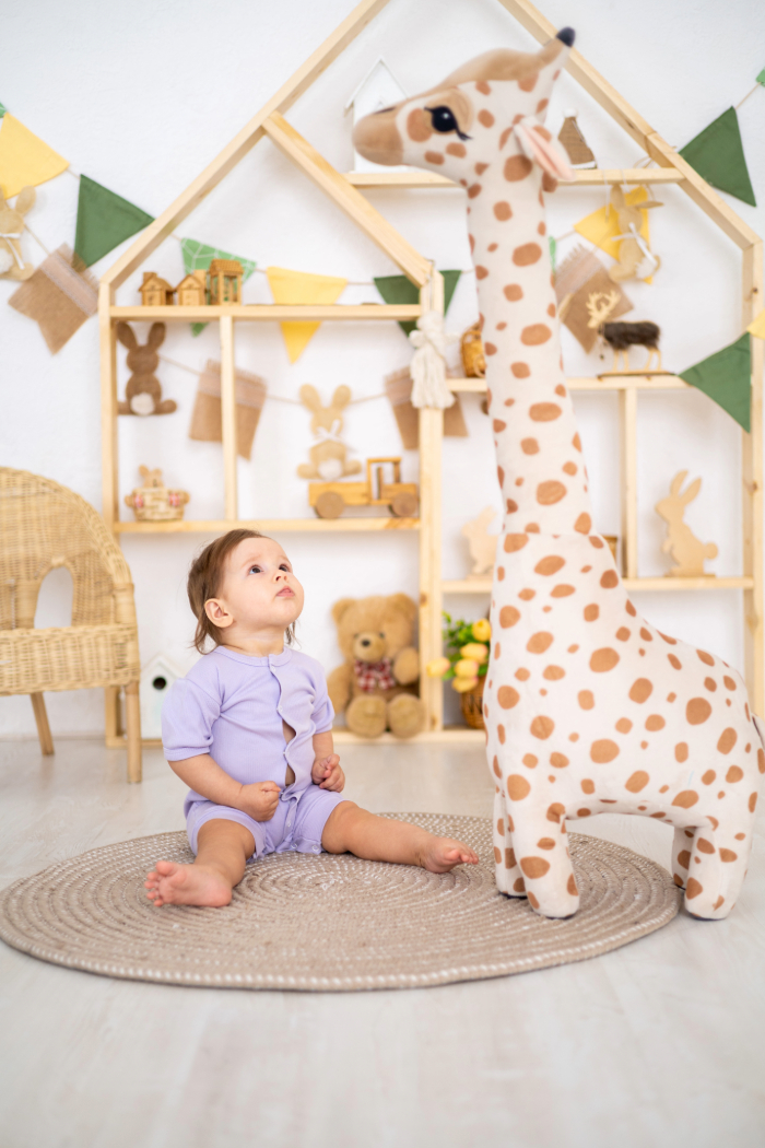 Bébé assis sur un tapis rond, regardant une grande peluche girafe, décor doux avec jouets en bois et guirlandes colorées.