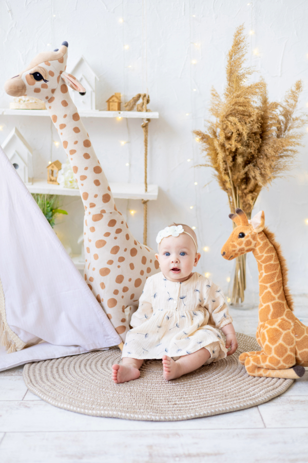 Bébé assis souriant sur tapis rond, entouré de deux grandes peluches girafes colorées.