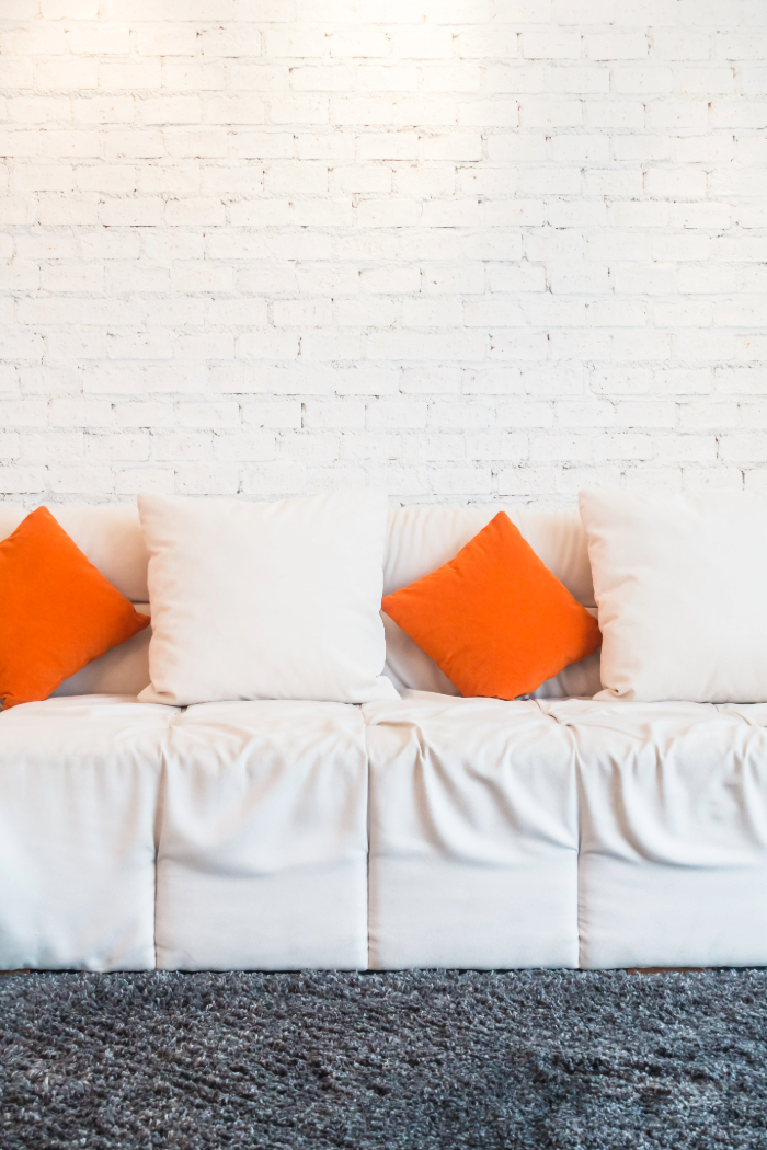 Un canapé blanc avec des coussins blancs et orange, sur un tapis gris et un mur en briques blanches.