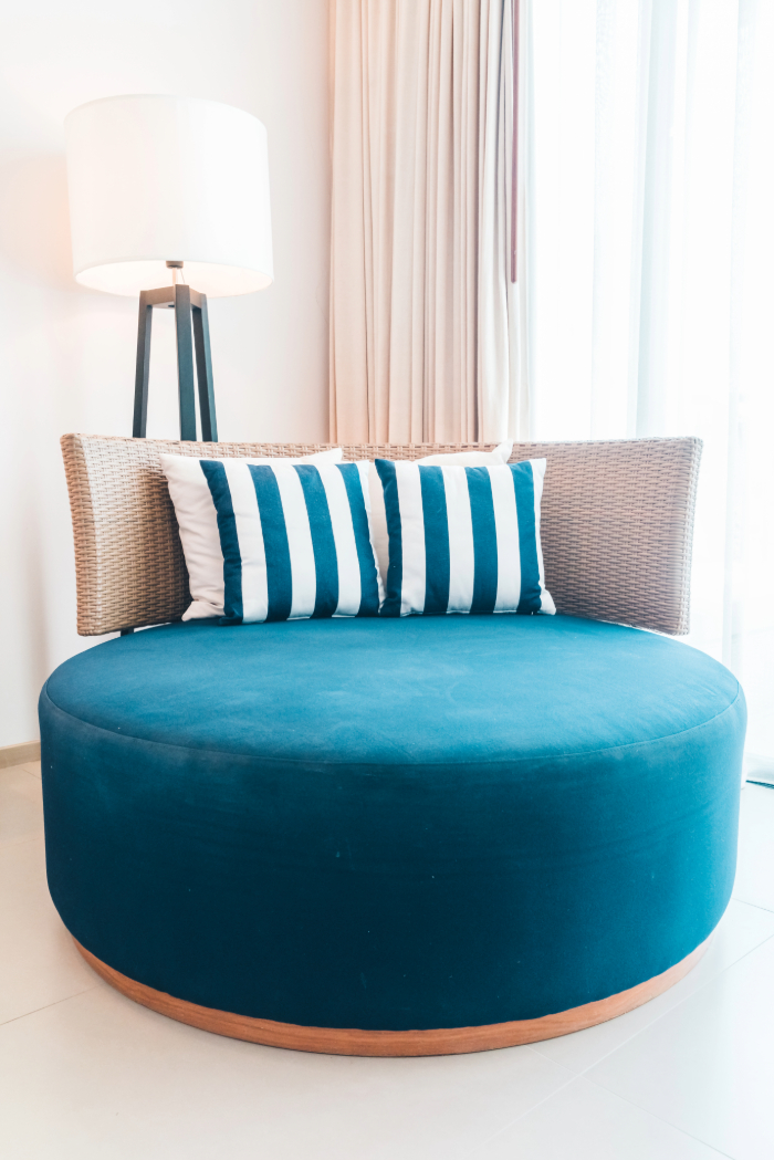 Un canapé rond en velours bleu, deux coussins rayés bleus et blancs, une lampe moderne sur pied, des rideaux clairs.