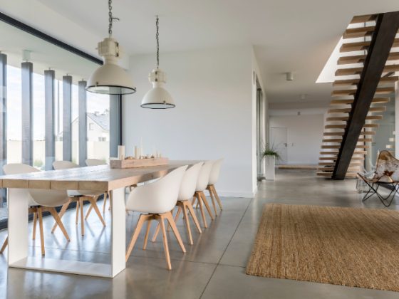 Intérieur scandinave avec table en bois clair, chaises blanches, suspensions industrielles, escalier flottant, tapis jute, fauteuil papillon et baie vitrée.