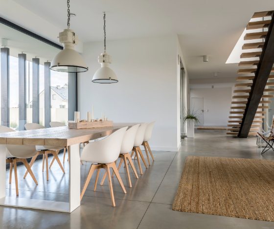 Intérieur scandinave avec table en bois clair, chaises blanches, suspensions industrielles, escalier flottant, tapis jute, fauteuil papillon et baie vitrée.
