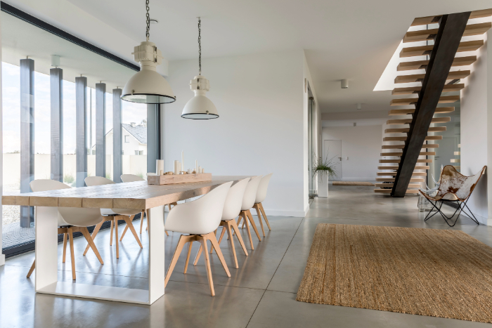 Intérieur scandinave avec table en bois clair, chaises blanches, suspensions industrielles, escalier flottant, tapis jute, fauteuil papillon et baie vitrée.