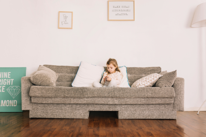 Une petite fille assise sur un canapé gris, entourée de coussins, dans un salon clair et minimaliste, calme.
