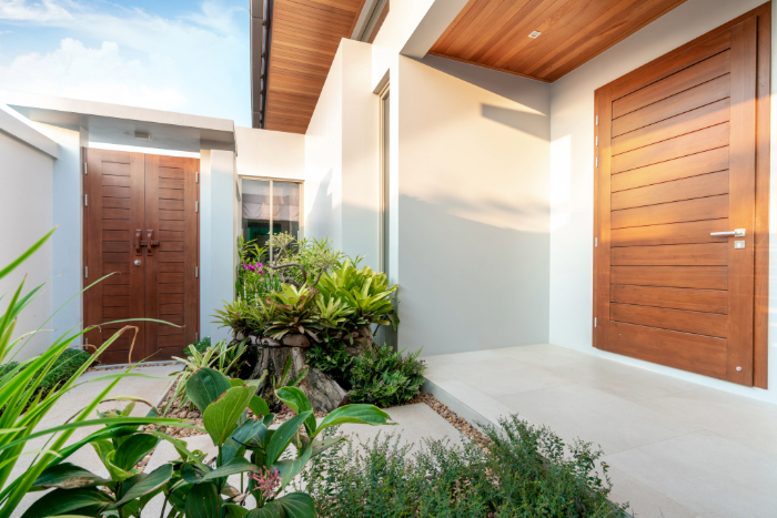 entrée maison moderne avec végétation luxuriante, murs blancs et porte en bois.