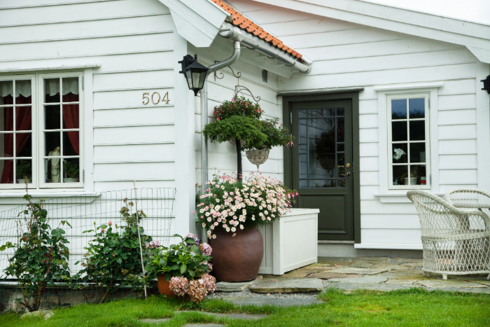 Aménagement extérieur devant une maison qui inclut des pots et jardinières fleuries, chaises en osier, petite pelouse verdoyante et une allée en pierres.