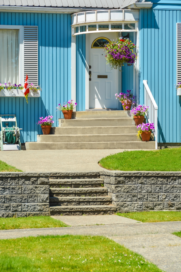 Aménagement extérieur devant maison incluant porte blanche, fleurs violettes en pots, escalier en béton et une pelouse verte.