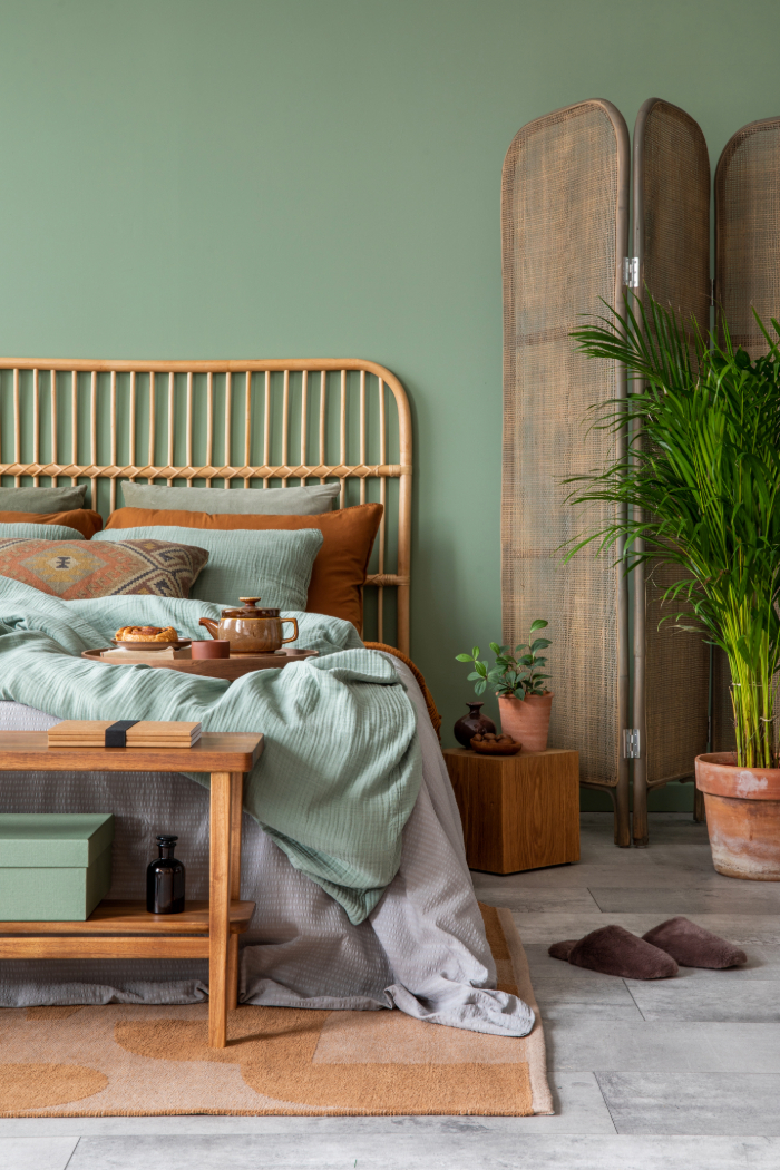 Chambre cosy et naturelle avec lit rotin, linge vert doux, coussins ocre, plantes, paravent, banc bois et tapis.