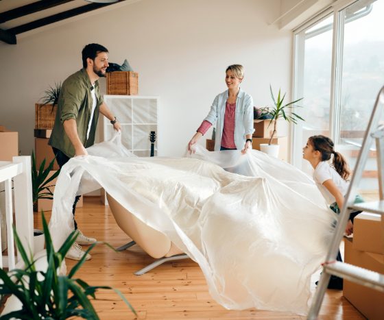 Une pièce lumineuse incluant un canapé, entouré de plantes et de cartons, avec trois personnes faisant la décoration de cette maison neuve.