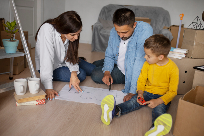 Une famille planifie un aménagement et une décoration d'une maison neuve, assise par terre avec des plans, entourée de cartons.