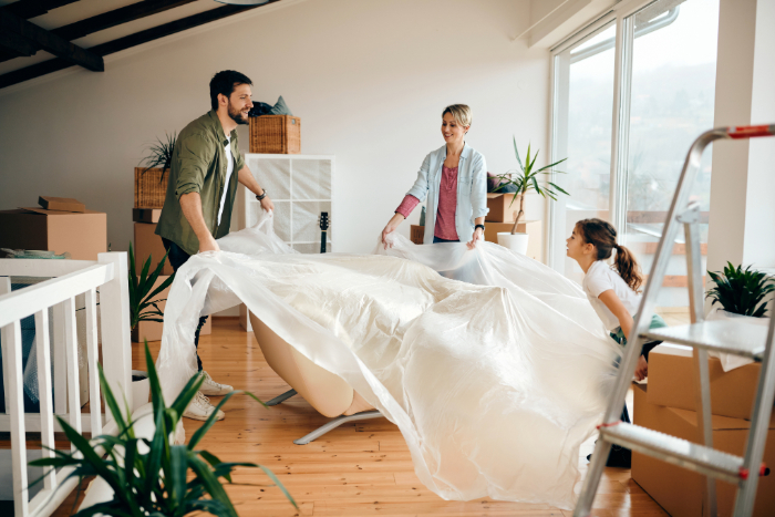 Une pièce lumineuse incluant un canapé, entouré de plantes et de cartons, avec trois personnes faisant la décoration de cette maison neuve.