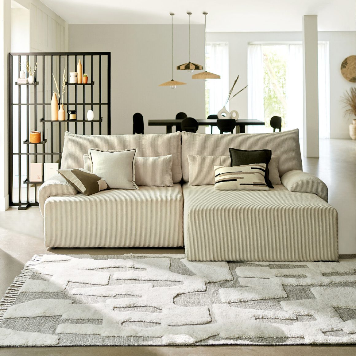 Salon moderne et lumineux avec canapé crème côtelé, coussins contrastés, tapis texturé et une étagère noire design.