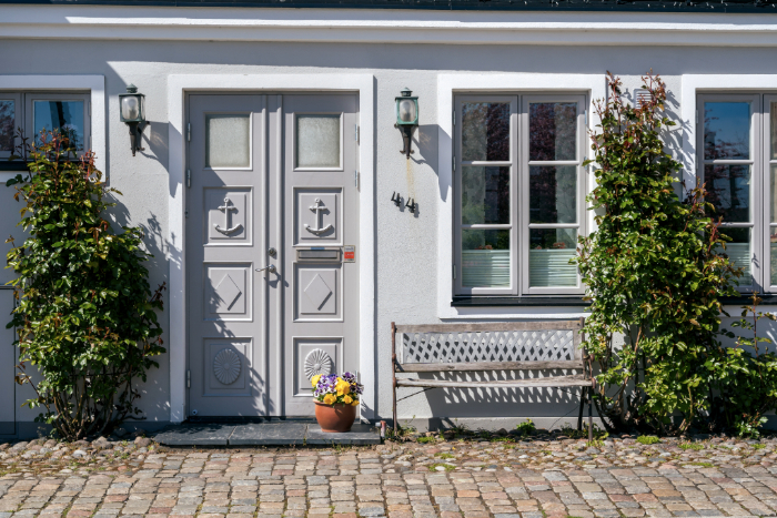Idée d'aménagement extérieur devant maison avec porte décorée d'ancres, fleurs en pot, banc, végétation et éclairages.