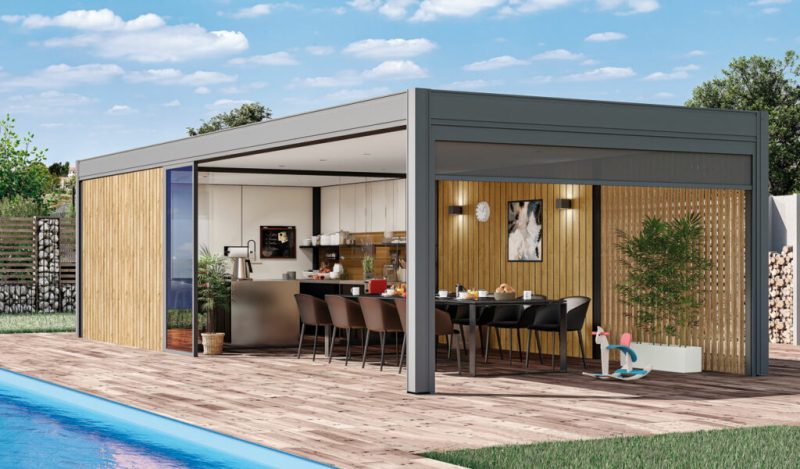 Pool house avec cuisine d'été incluant une grande table à manger noire, des chaises assorties, un bardage bois, une pergola grise, des plantes décoratives, un jouet enfant et un parquet en bois.