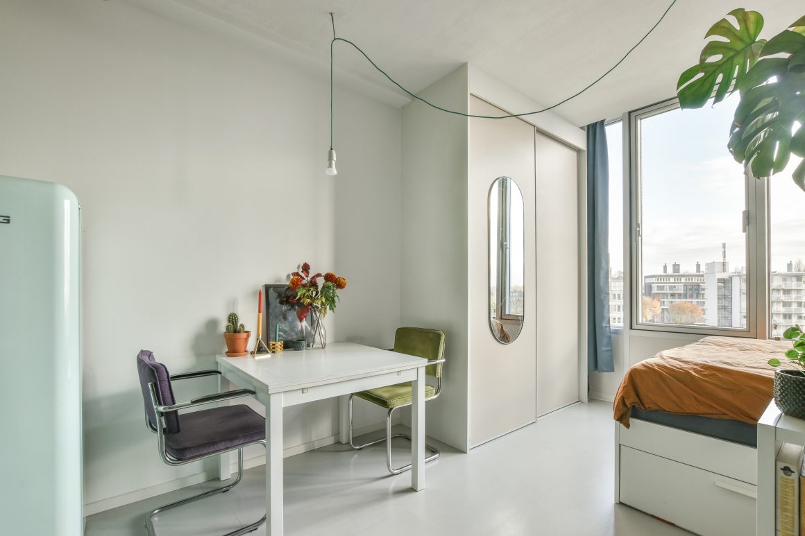 Petit espace lumineux avec table blanche, deux chaises colorées, plante, lit, armoire et grande fenêtre.