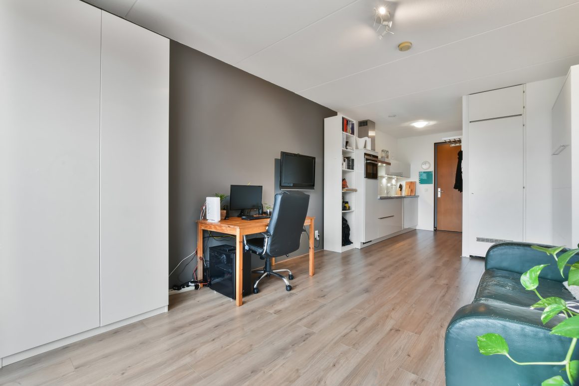 Aménagement studio 20m2 avec bureau en bois, chaise noire, canapé vert et cuisine blanche.