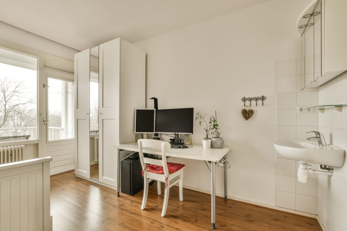 Pièce lumineuse avec bureau, deux écrans, chaise, armoire, plantes, lavabo et sol en bois clair.