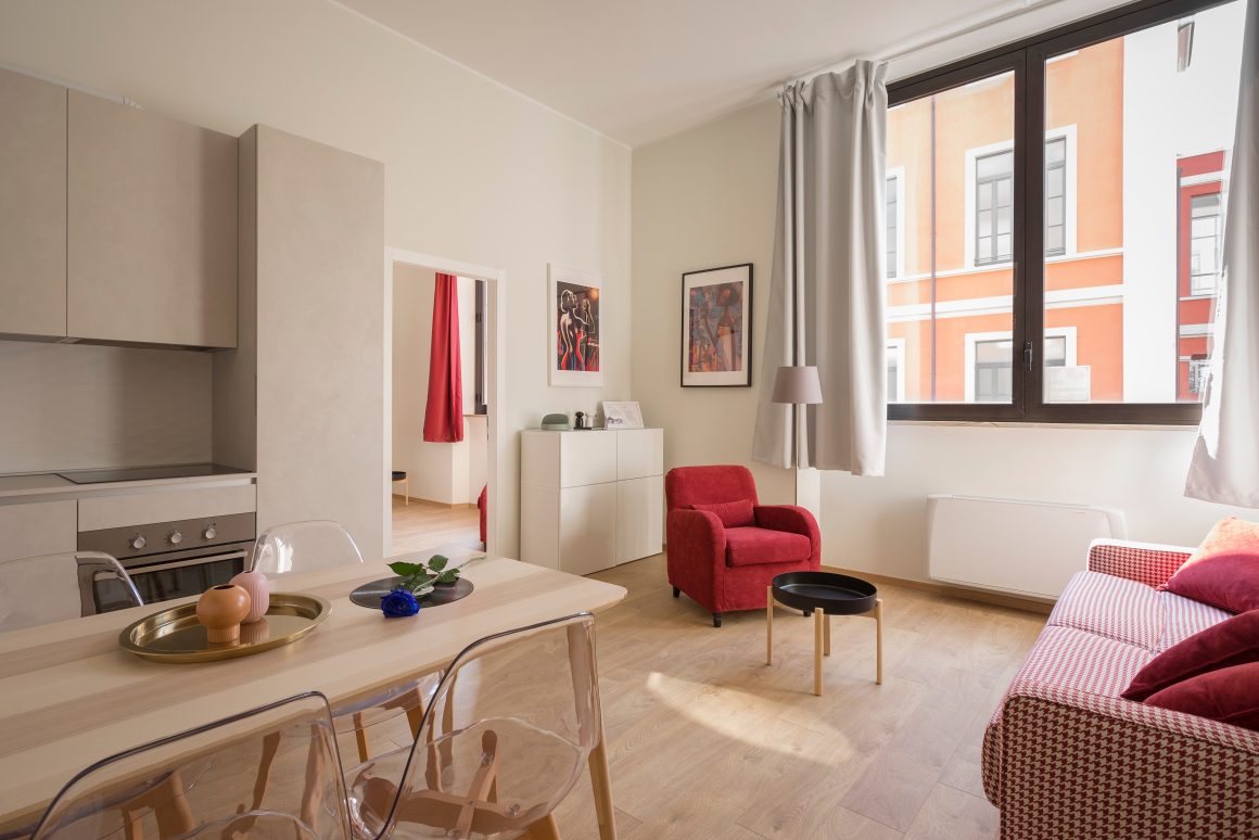 Décoration pour petit appartement, incluant un espace ouvert, avec des tons neutres clairs, rideaux blancs, tableaux, fauteuil rouge et table en bois.