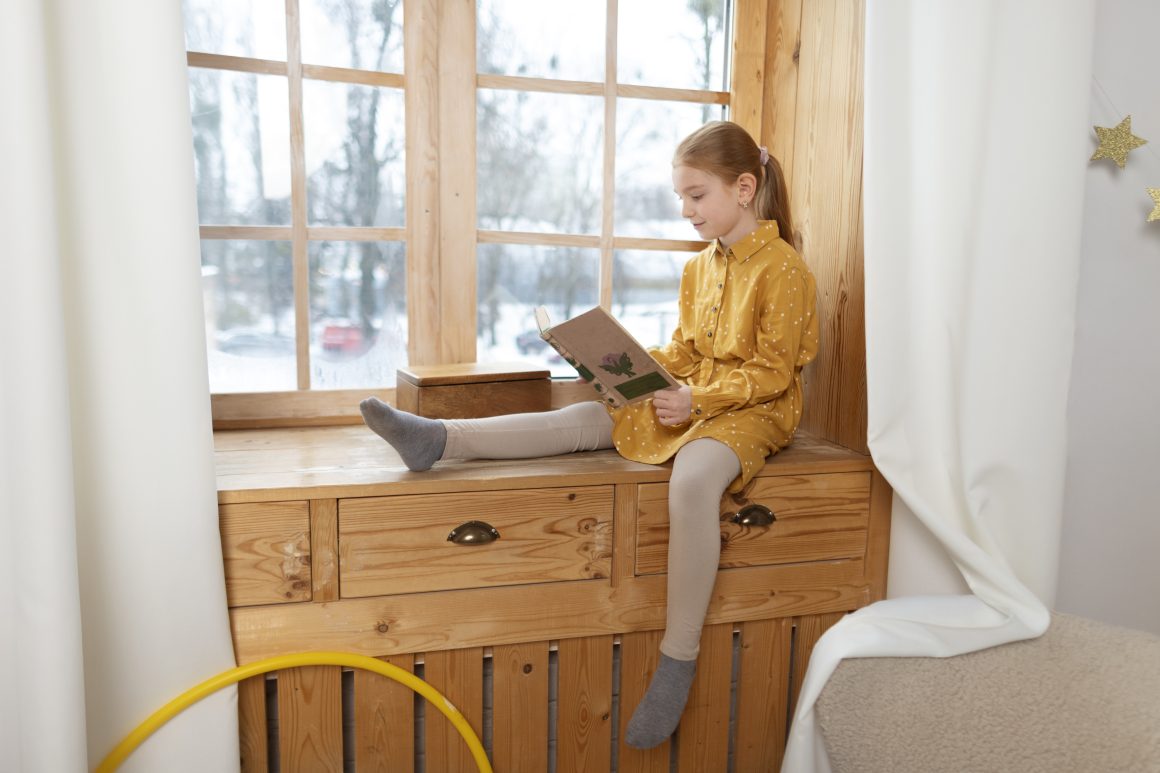 Une fille lit un livre assise sur meuble intégré dans le rebord de fenêtre.