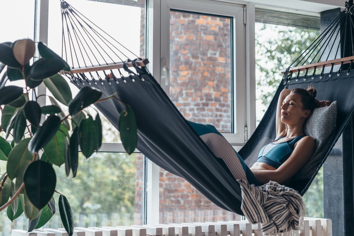Une femme détendue dans un hamac intérieur près d'une grande fenêtre, entourée de lumière naturelle et d'une plante.