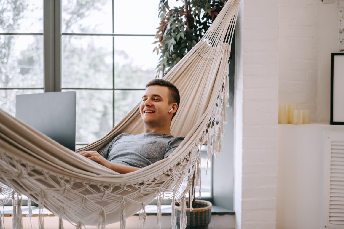 Un homme souriant travaille sur un ordinateur portable, allongé dans un hamac à l'intérieur.
