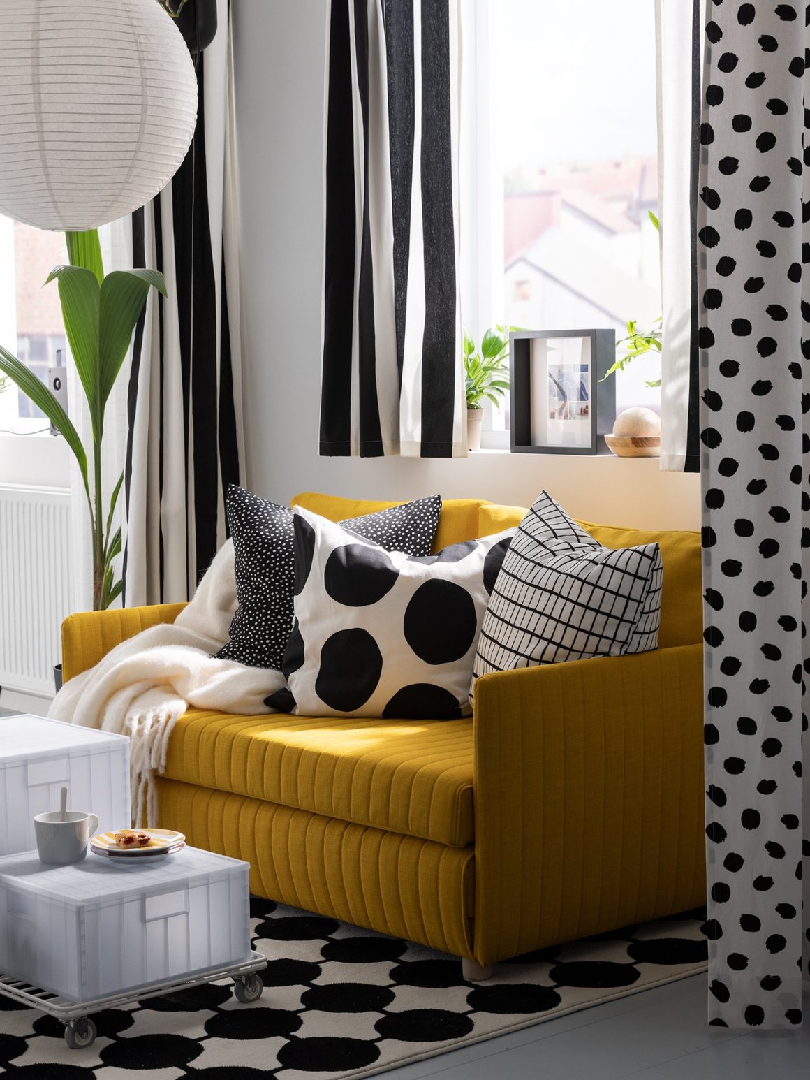 Canapé jaune vibrant avec coussins à motifs noirs et blancs avec rideaux rayés, décorant un petit appartement.