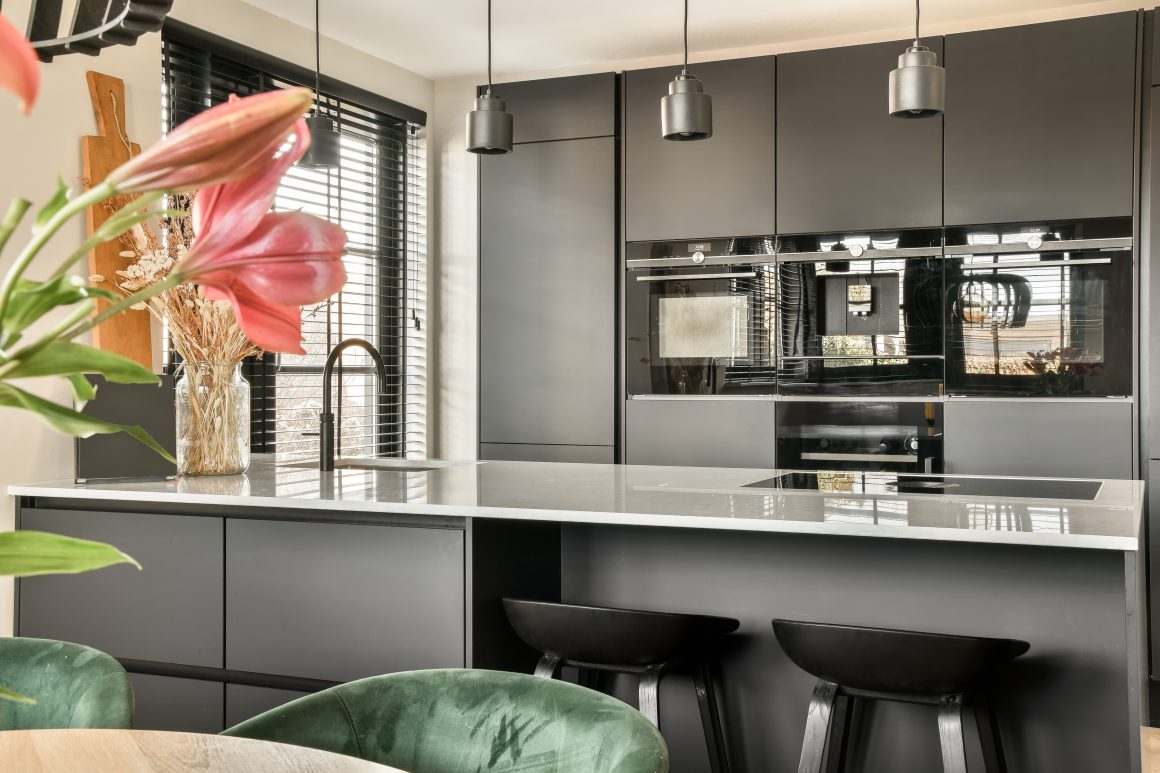 Modèle de cuisine moderne avec armoires gris foncé, îlot blanc, fenêtres à volets noirs et éclairage suspendu.