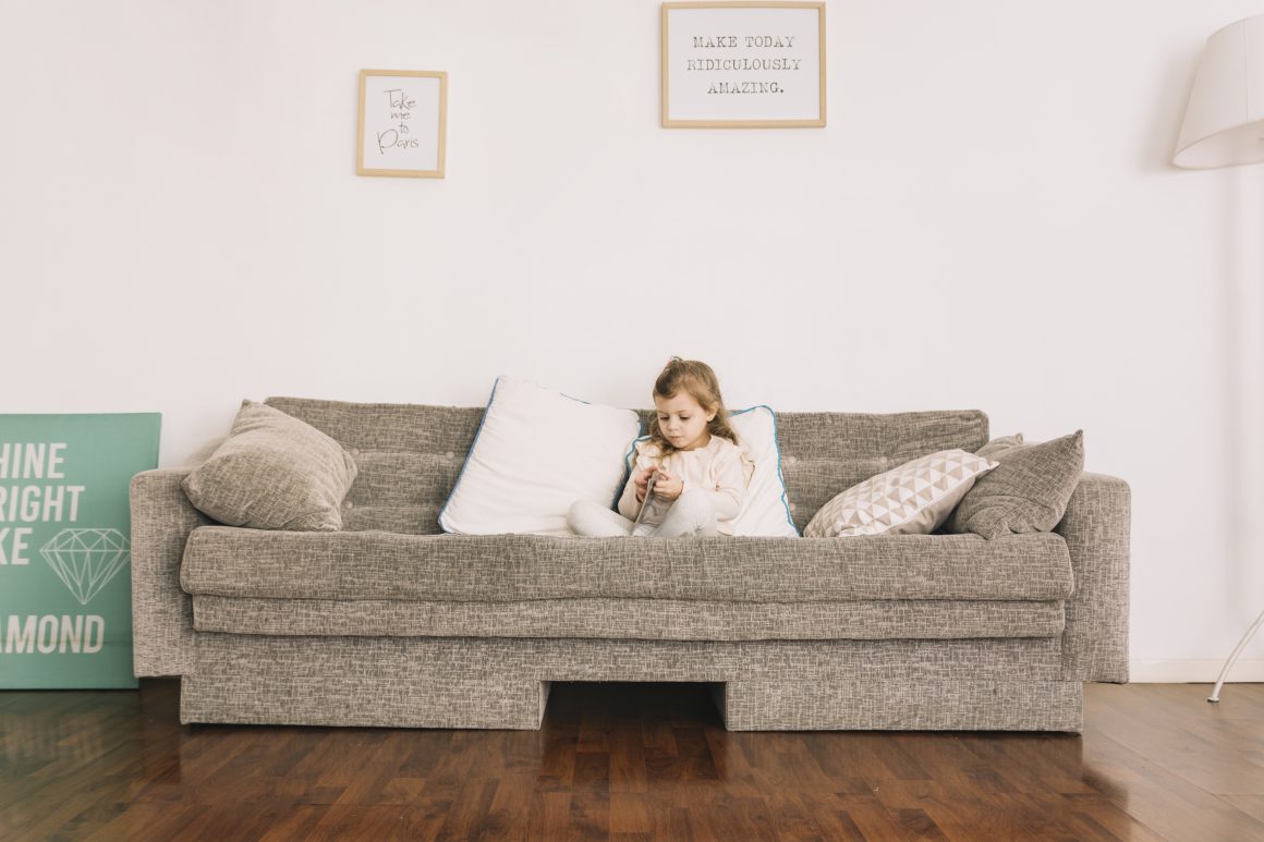 Une petite fille assise sur un canapé gris avec coussins, dans un salon lumineux au sol en parquet bois foncé.