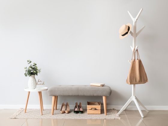 Entrée épurée avec portemanteau blanc, banc gris, table blanche, plante, chaussures et boîte en bois.
