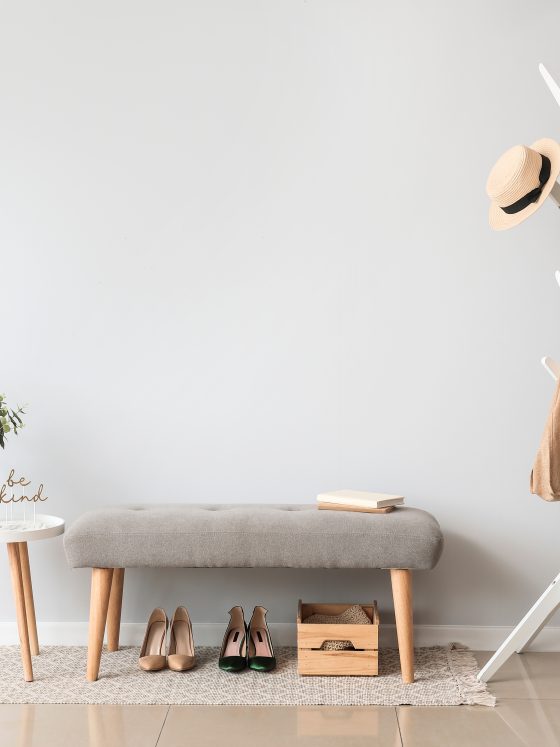 Entrée épurée avec portemanteau blanc, banc gris, table blanche, plante, chaussures et boîte en bois.