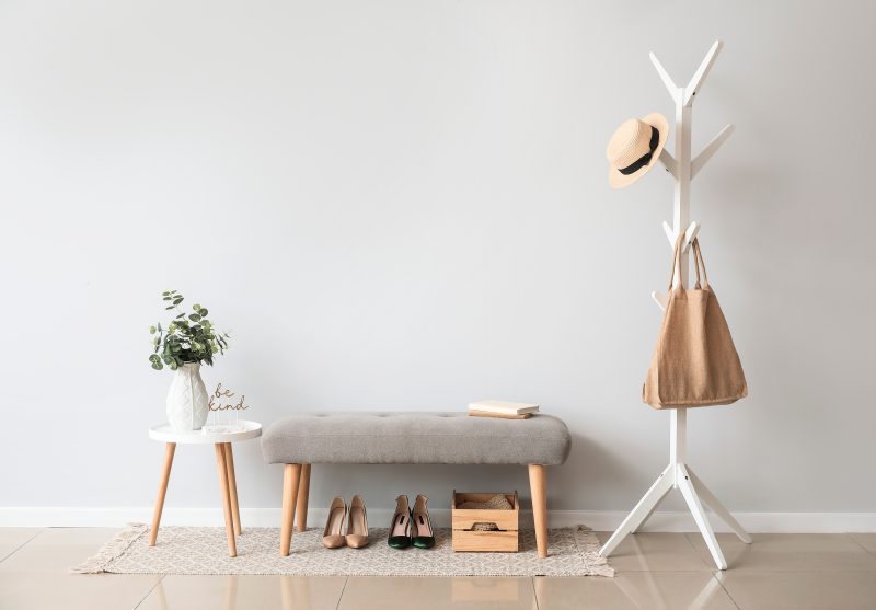 Entrée épurée avec portemanteau blanc, banc gris, table blanche, plante, chaussures et boîte en bois.