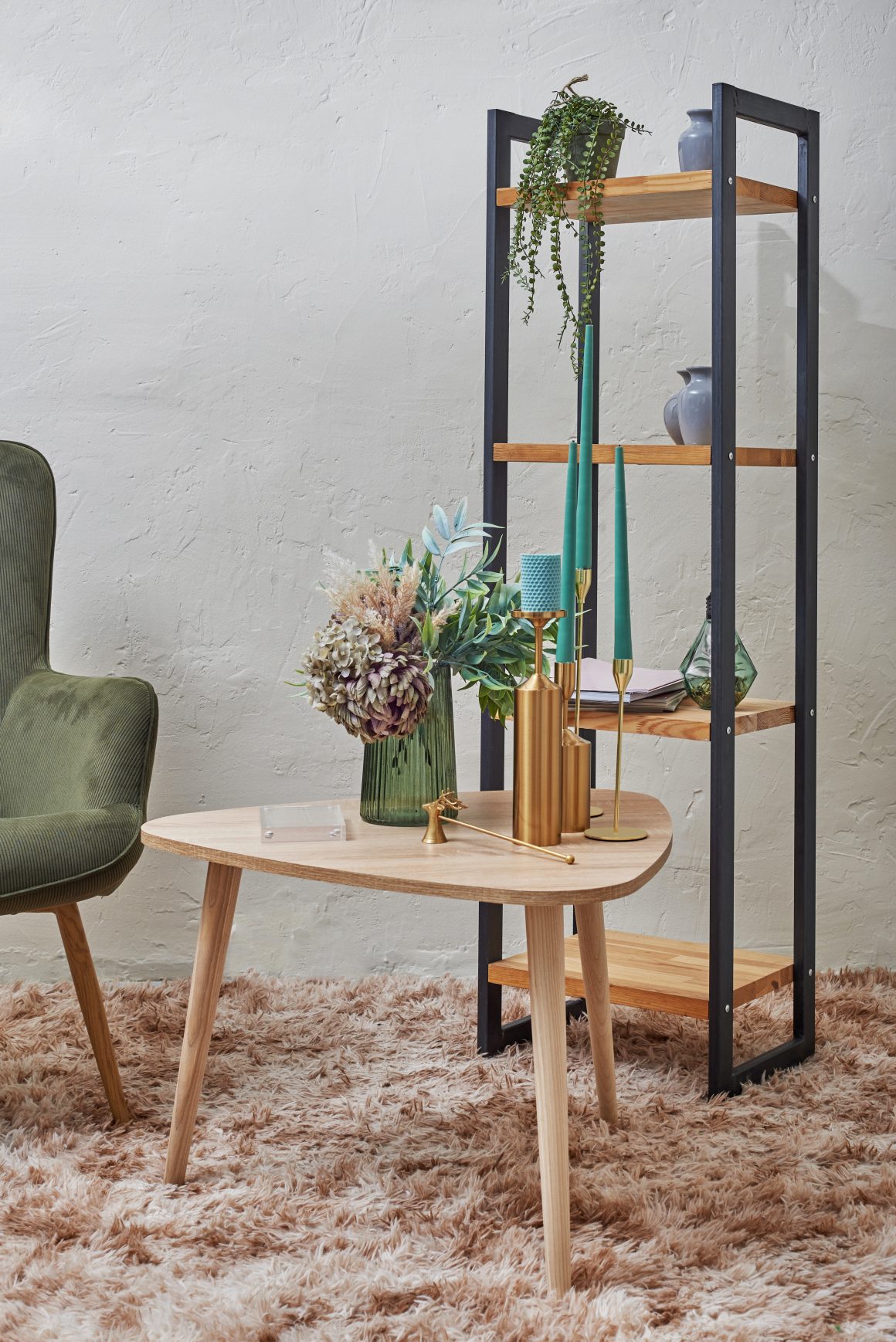 Table basse en bois avec bougeoirs dorés, bougies vertes, vase avec fleurs, étagère et fauteuil vert.