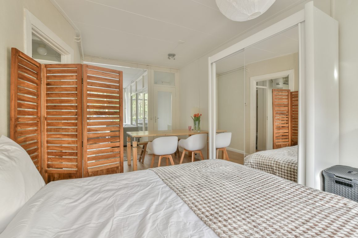 Chambre lumineuse avec lit, miroir, table, chaises, séparation en bois et déco simple.