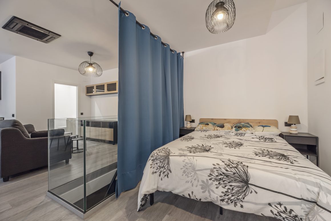 Chambre avec lit, rideau séparable, lampe suspendue et salon visible en arrière-plan.