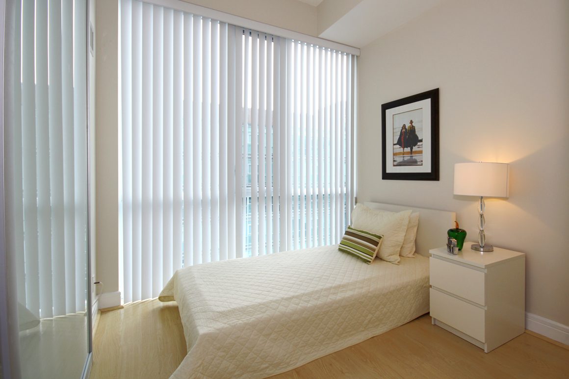 Chambre minimaliste incluant lit, lampe, tableau avec une grande fenêtre habillée par un store californien.