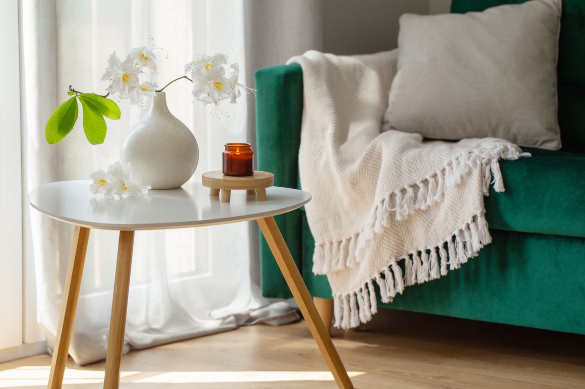 Table basse blanche, vase avec fleurs blanches, bougie, canapé vert, couverture beige et coussin gris.