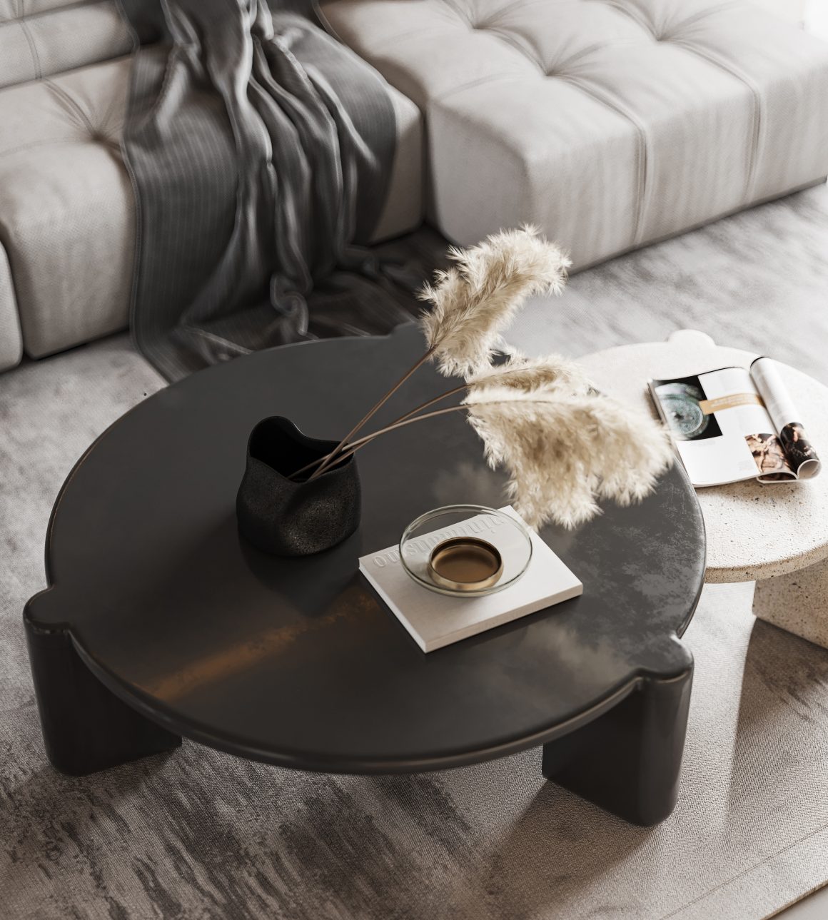 Table basse ronde noire avec vase, plume décorative, livre, tasse, canapé beige et plaid gris.