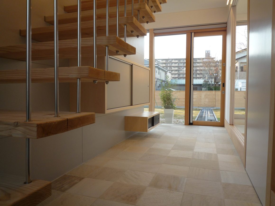 Intérieur lumineux avec escalier en bois, sol en pierre beige et grande porte-fenêtre.