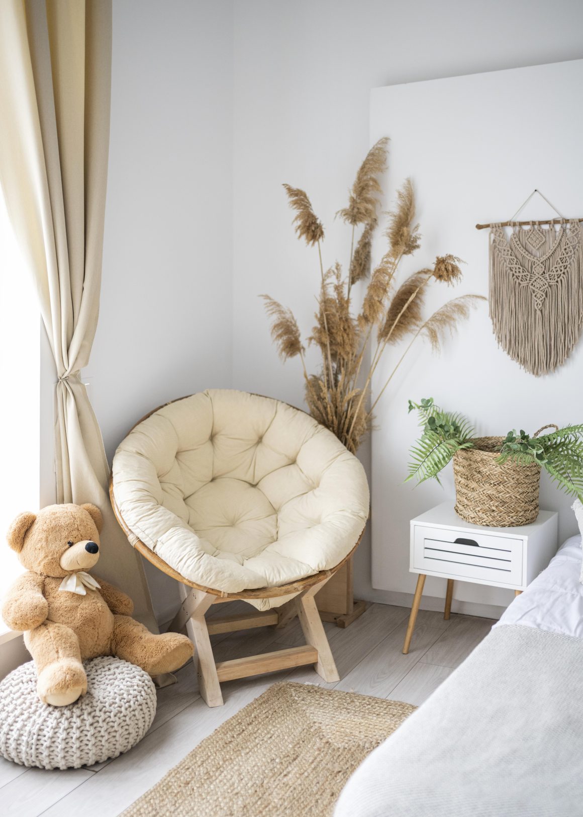Coin cosy avec fauteuil rond, peluche, rideaux beiges, panier tissé, plantes, tapis naturel et déco murale macramé.
