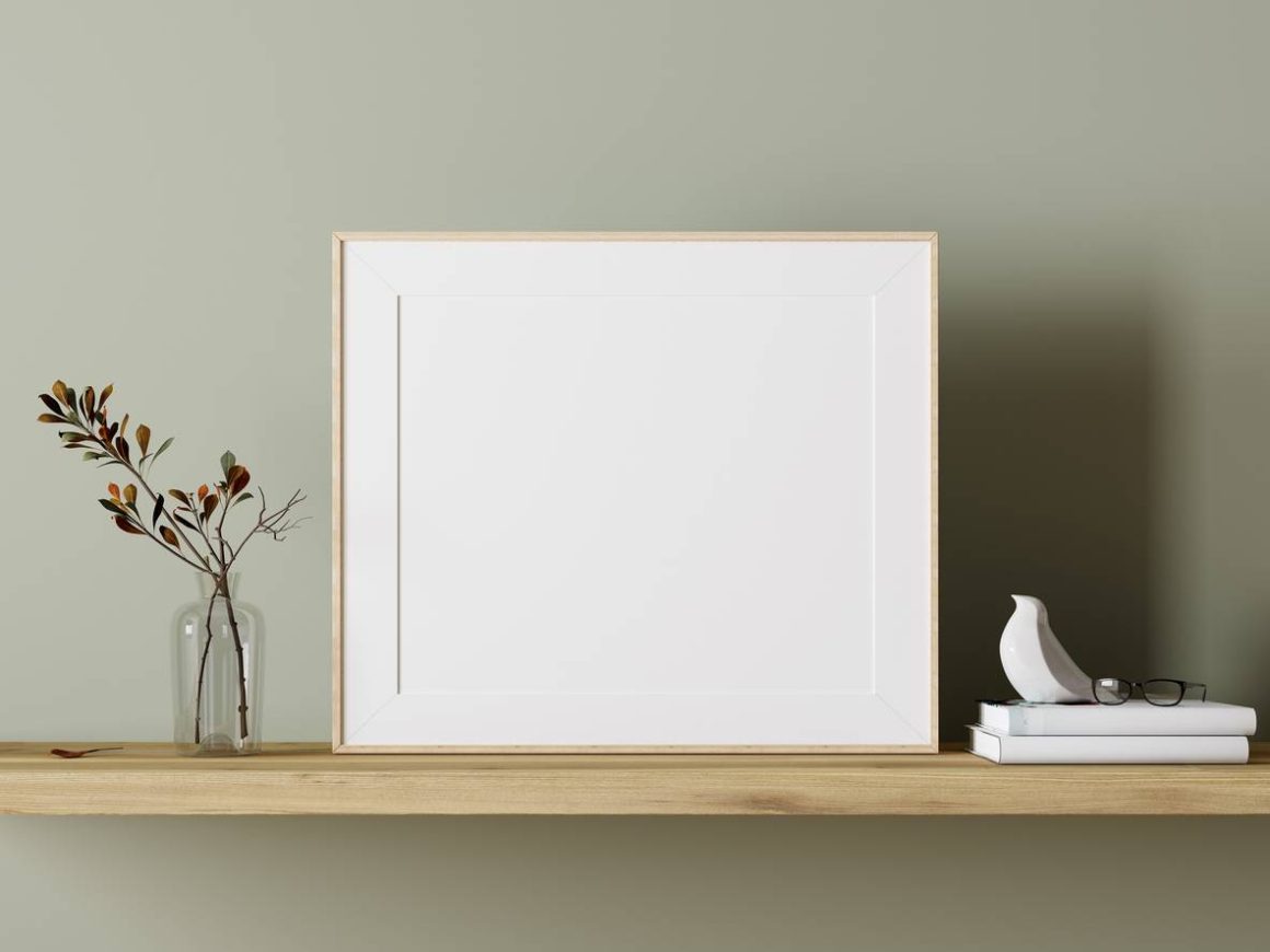 Cadre photo vide en bois clair, vase transparent avec branches, livres blancs, statue oiseau blanche et lunettes sur étagère en bois naturel.