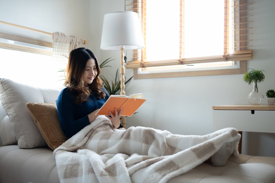 Une femme détendue lit un livre sur un canapé, sous une couverture douce, dans une pièce lumineuse et confortable.
