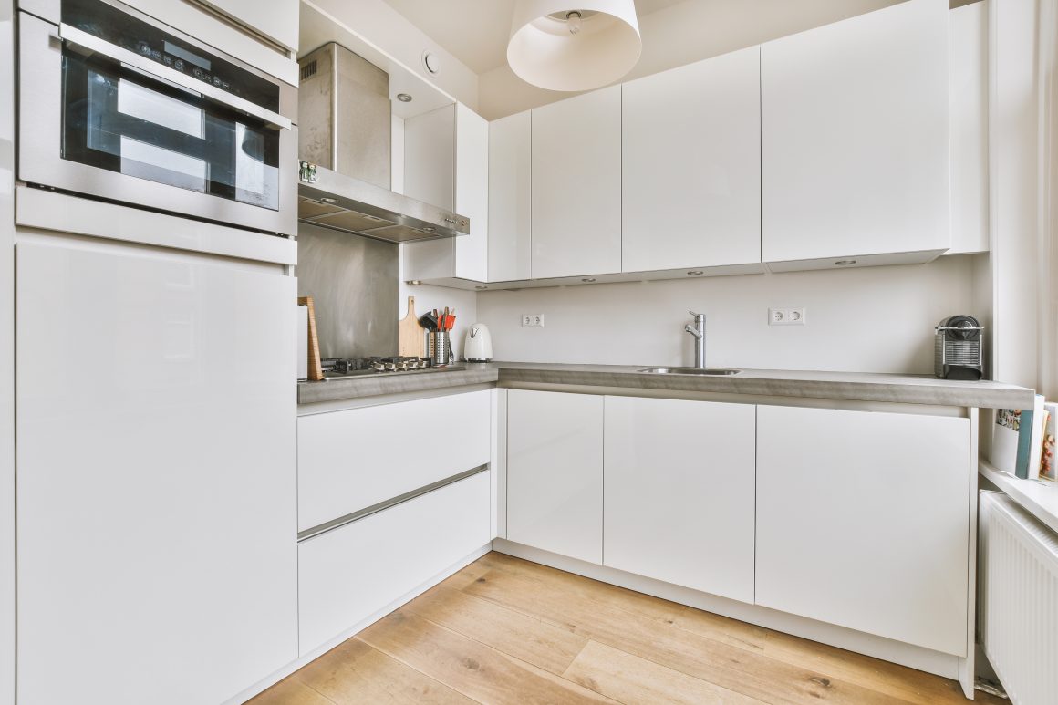 Cuisine blanche moderne avec armoires installées en haut et en bas, four intégré, hotte, plan de travail en béton, évier et sol en bois clair.